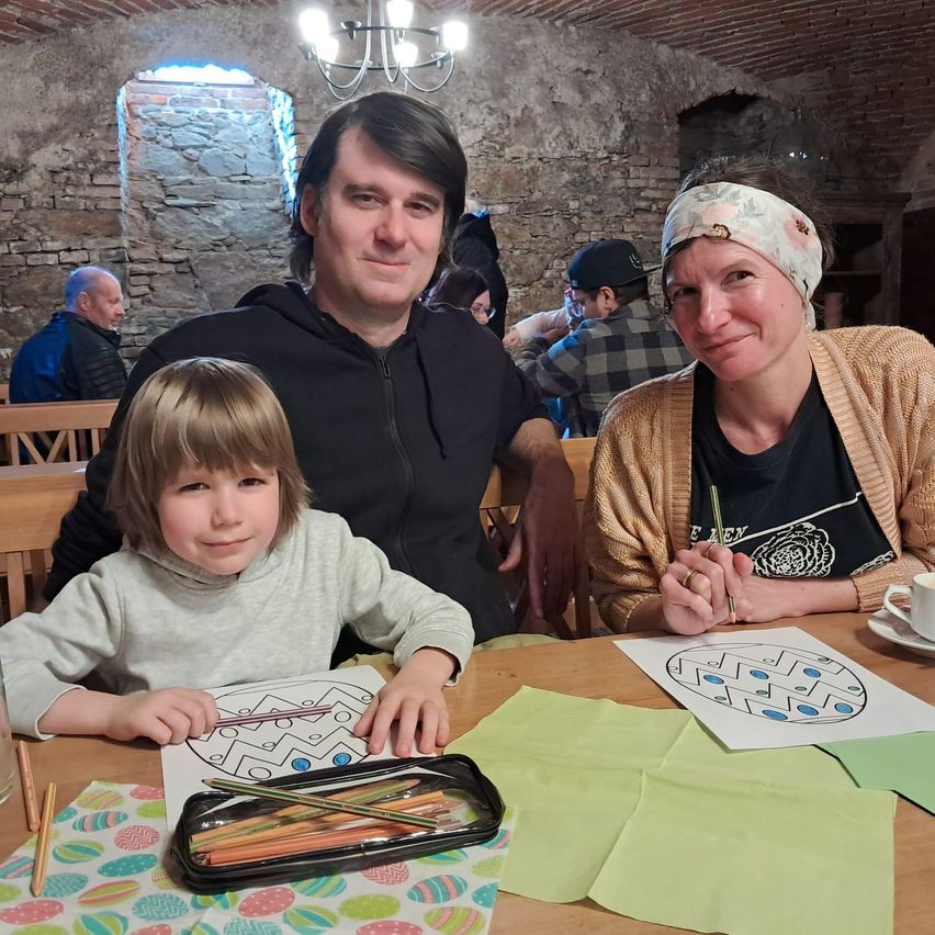 A family of three sits at a table, with a young girl coloring a maze. The man and woman wear casual clothing. Behind them, other people sit and talk, with a chandelier hanging from the ceiling.