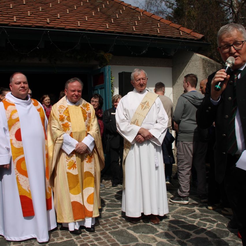 Three priests in white robes stand outside a building with a microphone. One holds a microphone, and the others stand behind him.