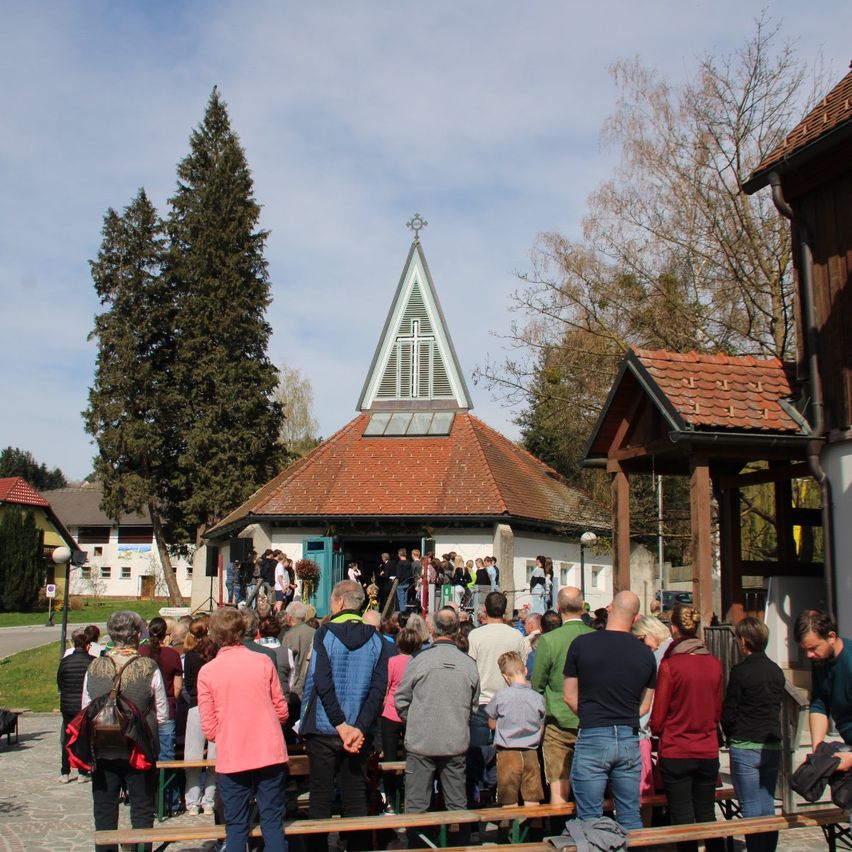 A group of people gather outside a chapel with a cross on its roof. They stand and sit on benches, while others are entering the chapel. Trees and houses are in the background.