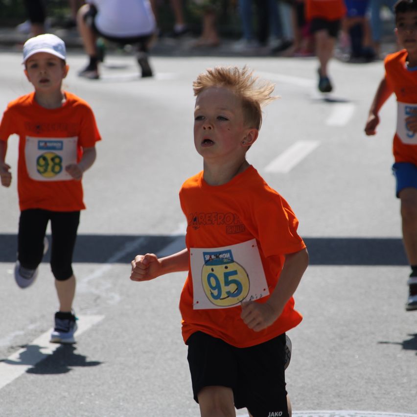 Drei Jungen in orangefarbenen Shirts und Shorts laufen in einem Marathon. Der Junge vorne hat die Nummer 95 auf seinem Shirt.