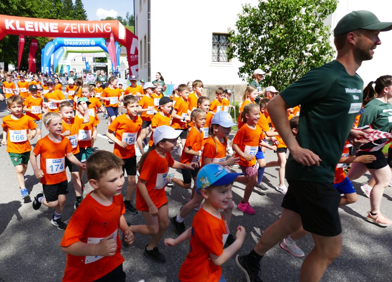 Eine Gruppe von Kindern in orangefarbenen Shirts mit Nummern nimmt an einem Rennen teil. Zuschauer und ein Baum säumen die Straße. Ein Mann in Grün geht nebenher.