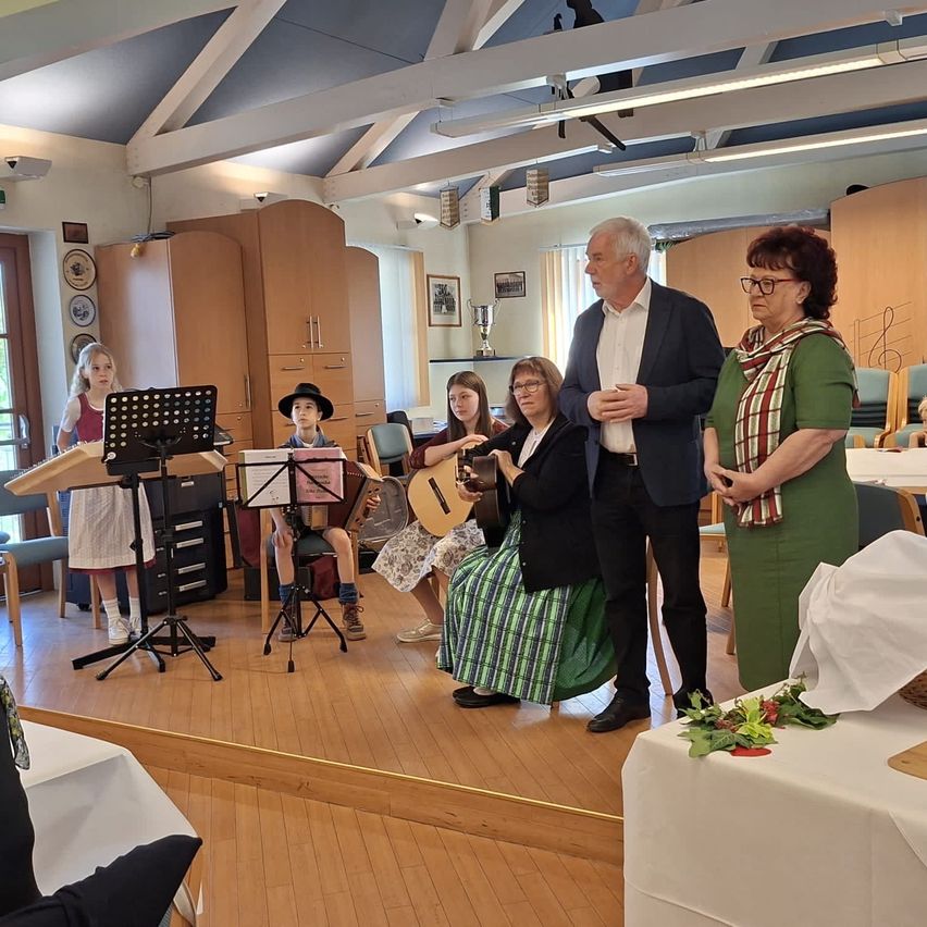 Eine Gruppe von Menschen in einem Raum, ein Mann steht vor ihnen, andere halten Gitarren. Ein Tisch mit Blumen und ein Pokal ist in der Nähe.
