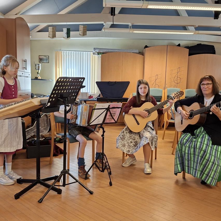 Vier Personen spielen in einem Raum Musikinstrumente. Zwei sitzen auf Stühlen und spielen Gitarre, während die anderen beiden stehen, einer spielt Akkordeon und der andere ein Holzinstrument.