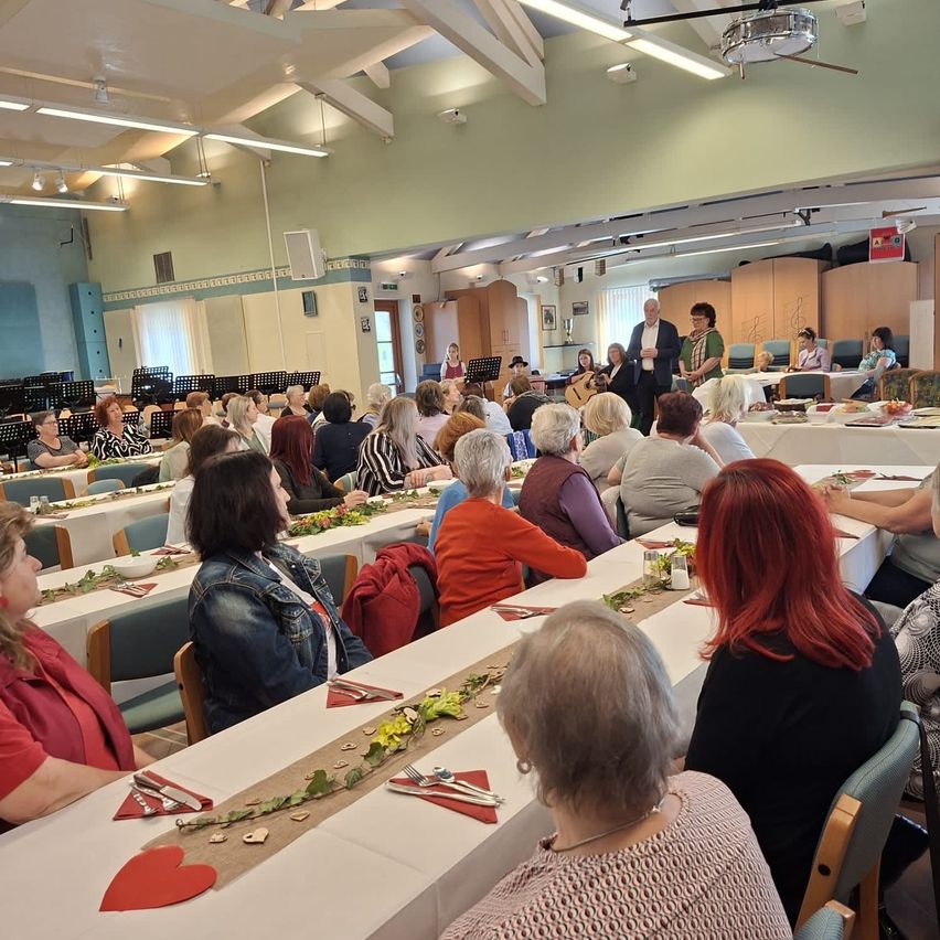 Eine große Gruppe von Menschen sitzt an langen Tischen in einem Raum, einige sitzen im Hintergrund. Eine Person steht vor den Tischen und einige andere stehen dahinter. Die Tische sind mit Blumen geschmückt und auf den Tischen stehen Musikinstrumente.