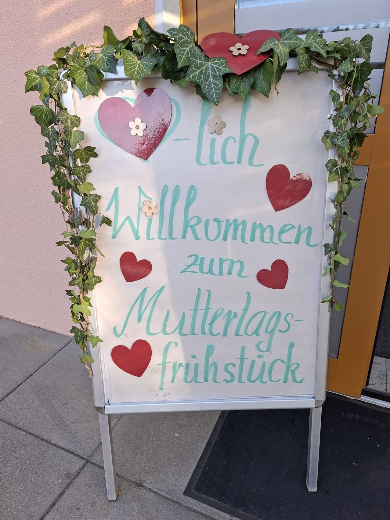 Ein weisses Schild mit Efeu und Herzen, das 'Herzlich Willkommen zum Muttertagsfruhstuck' in Blau und Rot besagt, ist an einem Metallständer befestigt.