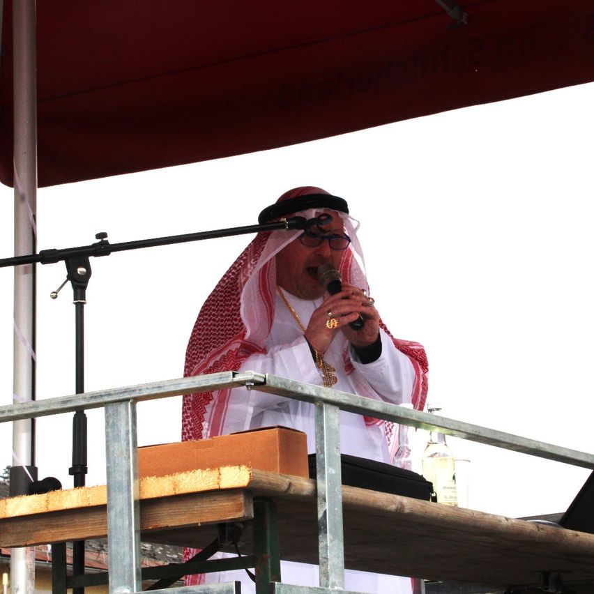 Ein Mann mit Brille und Keffiyeh spricht in ein Mikrofon auf einer Bühne unter einem roten Baldachin.