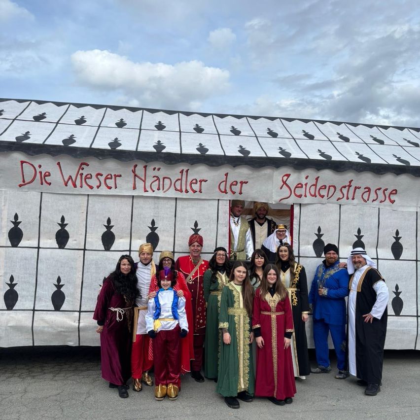 Eine Gruppe von Menschen in farbenfrohen Kostümen posiert vor einer dekorierten Bühne, auf der 'Die Wiser Händler der Seidenstrasse' steht. Der Hintergrund zeigt ein gemustertes Design.