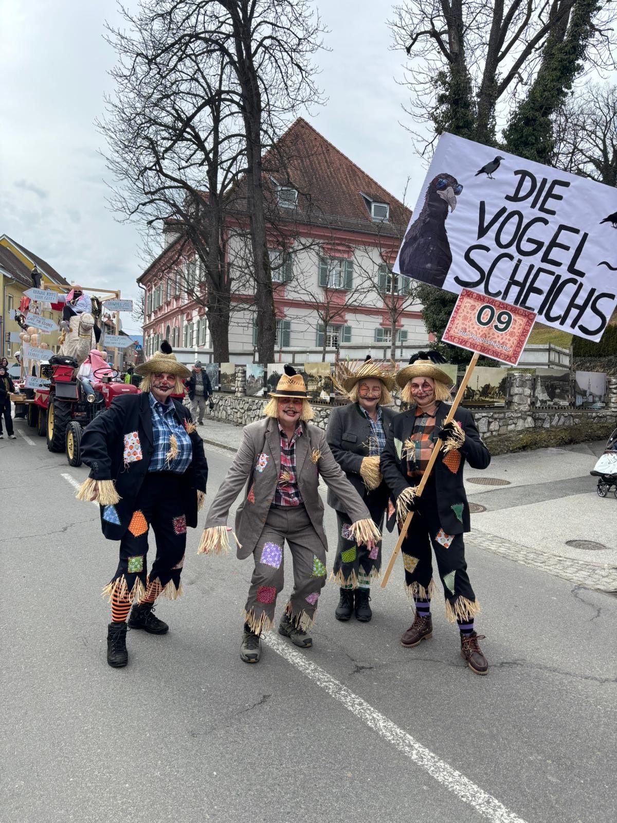 Drei Personen in Strohmannkostümen stehen auf einer Straße und halten ein Schild mit der Aufschrift 'Die Vogel Scheichs' und ein weiteres mit '09'. Hinter ihnen befinden sich Gebäude, ein Baum und ein Traktor. Der Himmel ist bedeckt.