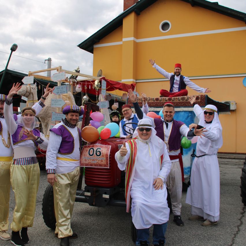 Eine Gruppe von Menschen in traditionellen Kostümen posiert vor einem dekorierten Traktor mit Luftballons, eine Person sitzt auf der Rückseite, vor einem gelben Gebäude.