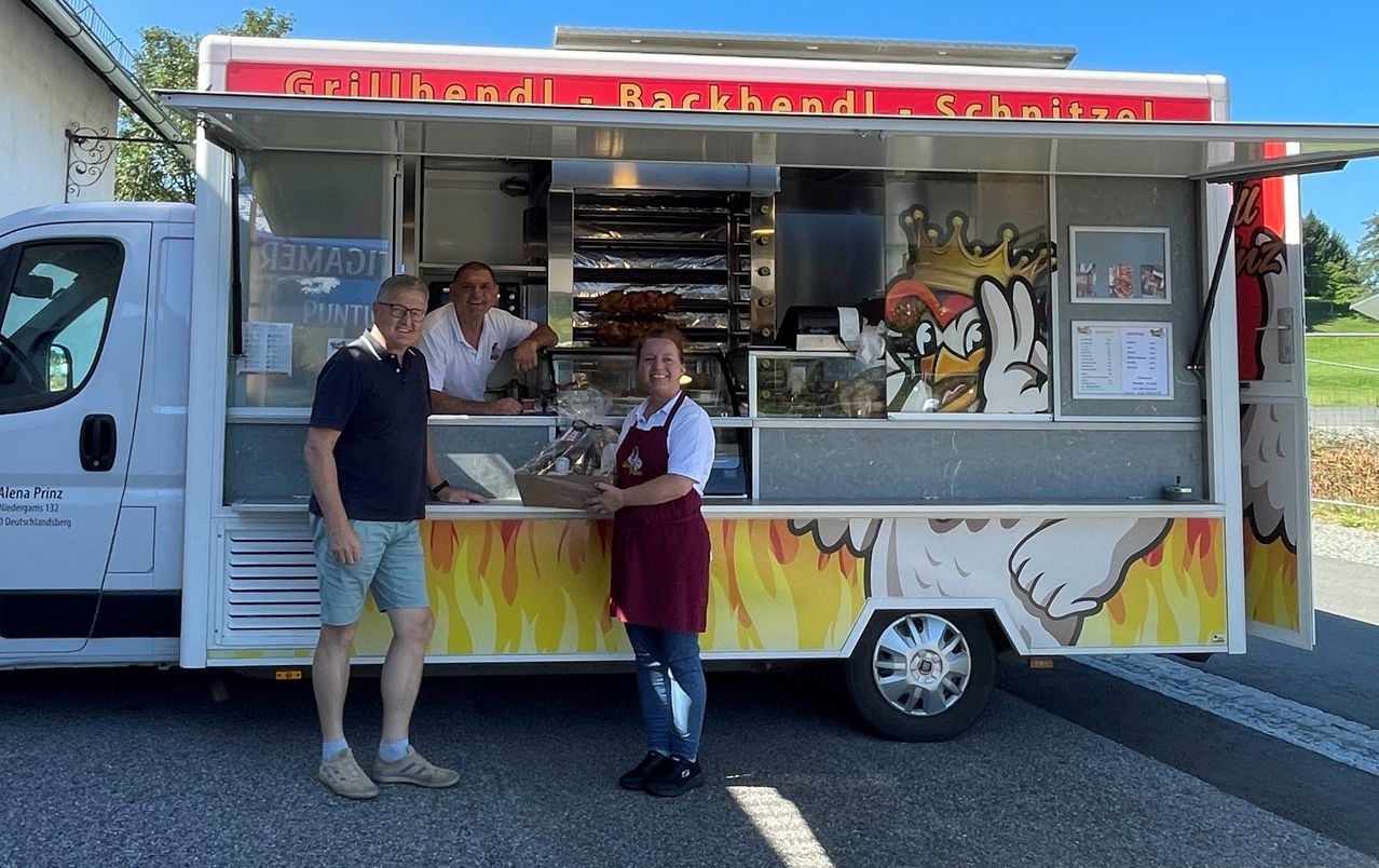 Drei Personen stehen vor einem Imbisswagen, zwei von ihnen lächeln. Der Mann auf der linken Seite trägt eine Brille und eine Uhr. Die Frau hält einen Korb mit Essen. Der Wagen hat ein farbenfrohes Design.