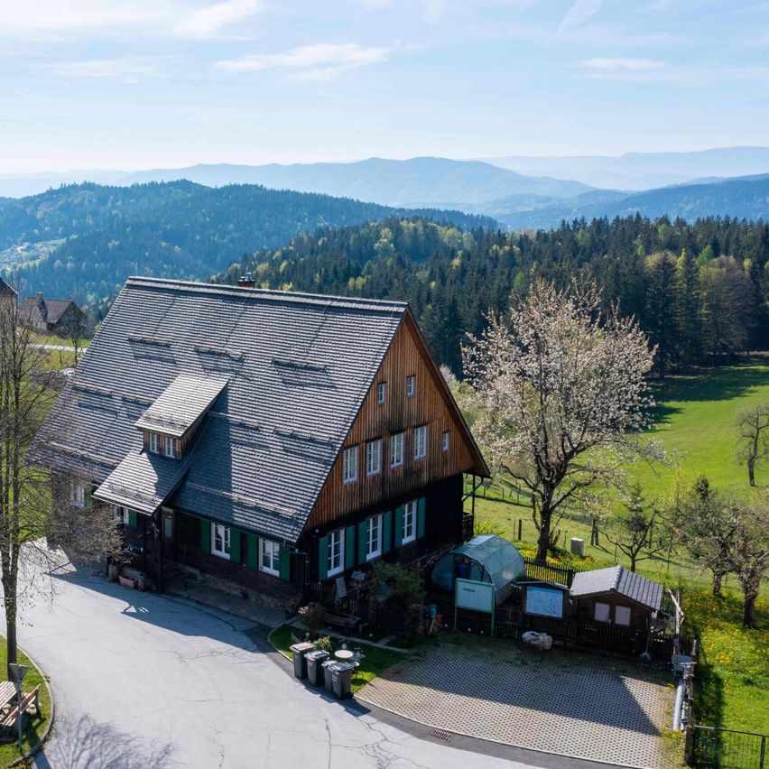 Eine Draufsicht auf ein rustikales Haus in einem Tal, umgeben von üppigen grünen Feldern und Bergen in der Ferne. Das Haus hat ein graues Dach, grüne Fensterläden und einen kleinen Garten.