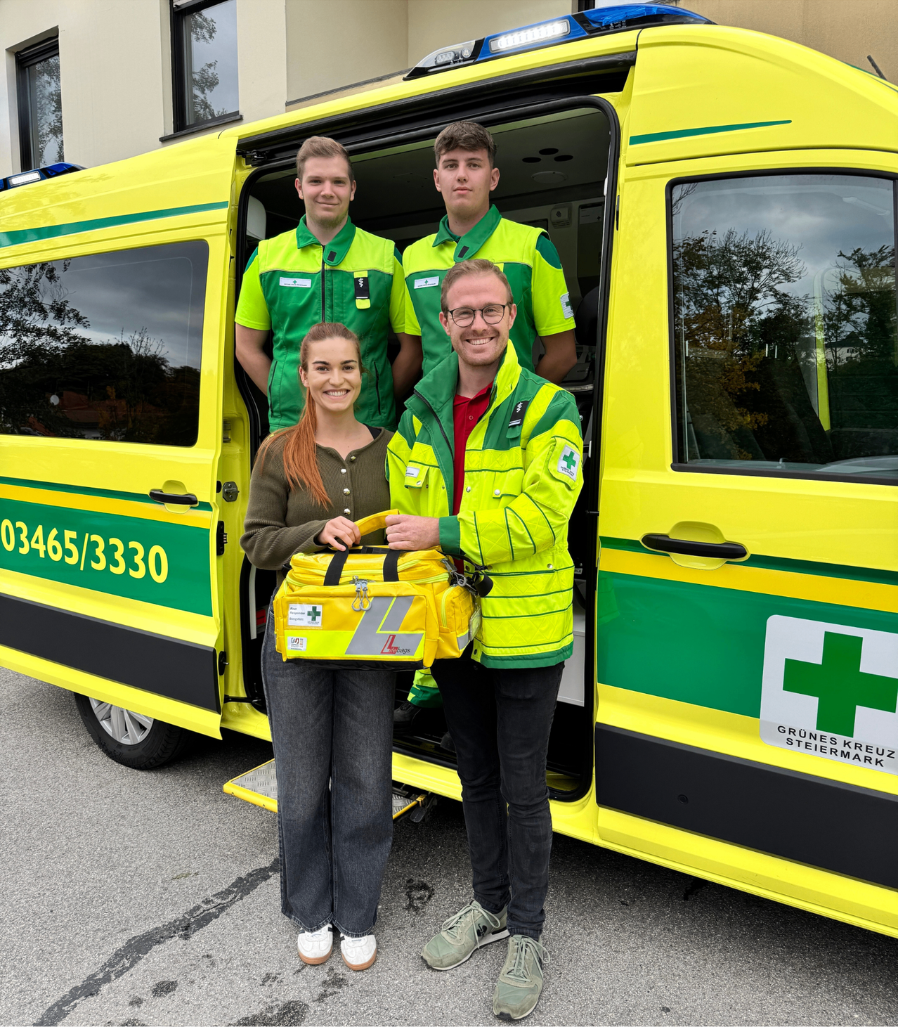 Vier Personen stehen vor einem Krankenwagen, zwei Männer in grünen Westen im Fahrzeug, eine Frau mit einer gelben Tasche und ein Mann mit Brille lächelt.