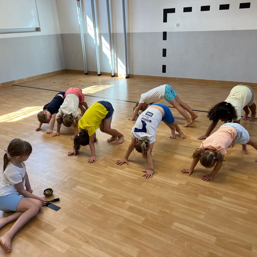 Eine Gruppe von Kindern übt Yoga in einer Turnhalle. Sie machen verschiedene Posen, einschließlich einer Handstand. Der Raum hat Holzböden und weiße Wände.