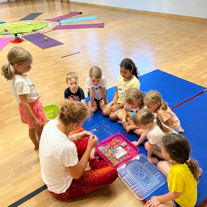 Eine Gruppe von Kindern und einer erwachsenen Frau sitzen auf einer blauen Matte und beschäftigen sich mit einer Aktivität. Die Frau hält eine Schale mit Perlen und die Kinder beobachten sie. Der Raum hat Holzböden und Yogamatten auf dem Boden.