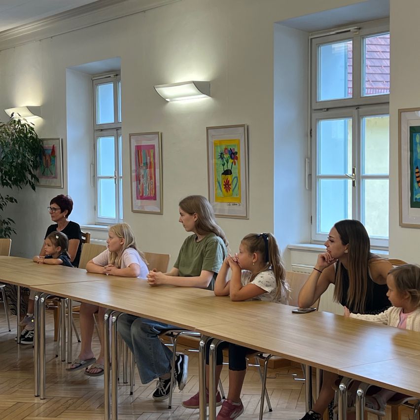 Eine Gruppe von Frauen und Kindern sitzt an einem langen Tisch in einem Raum mit großen Fenstern und Gemälden an der Wand.