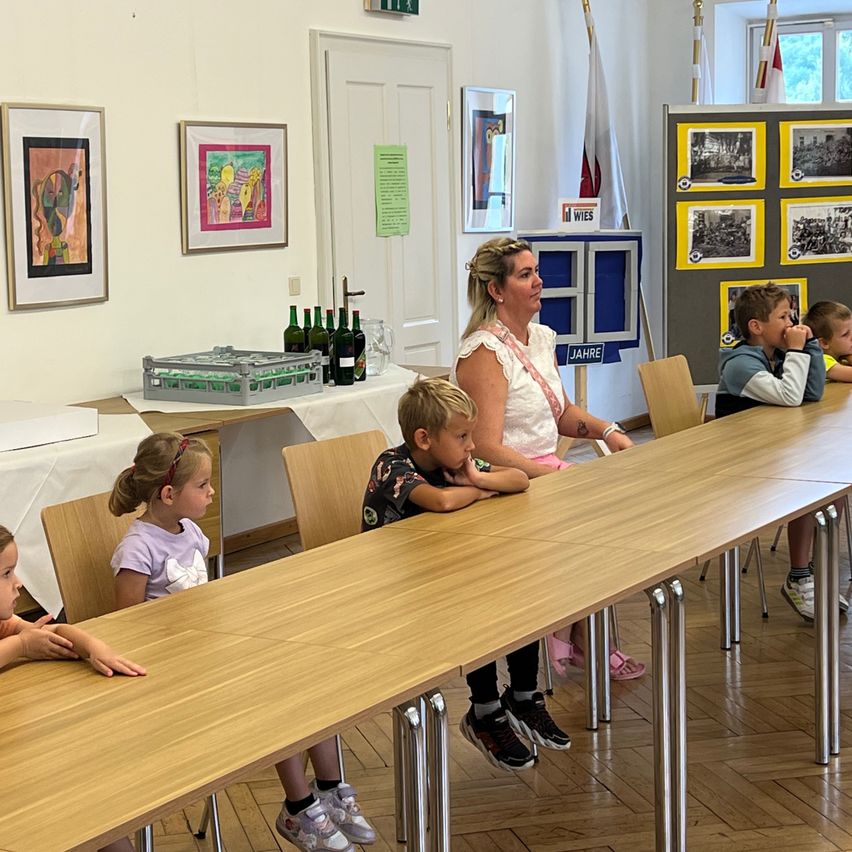 Eine Gruppe von Kindern und einer erwachsenen Frau sitzen an einem langen Holztisch in einem Raum mit gerahmten Bildern an der Wand und einem Tisch mit Flaschen.