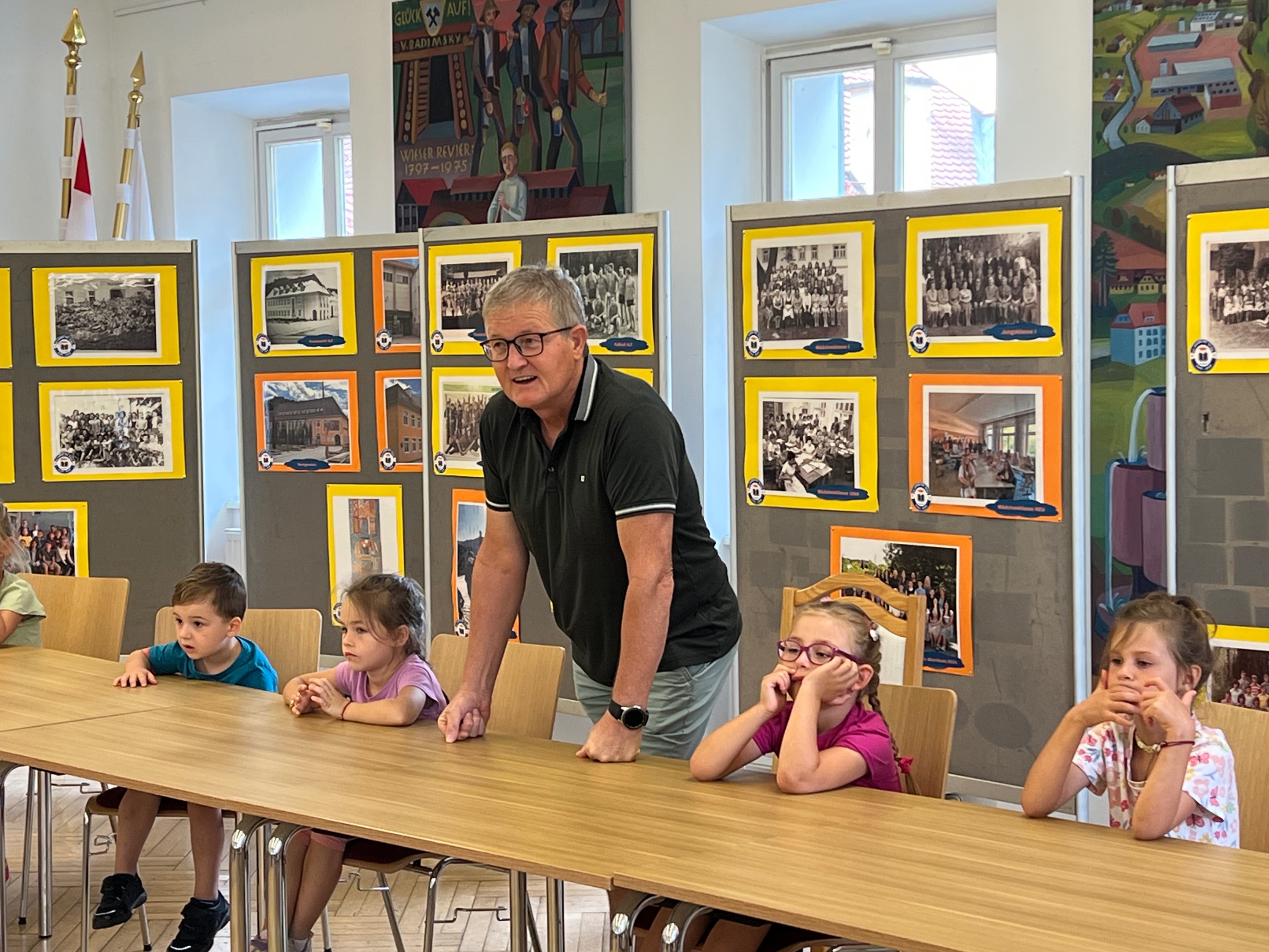 Mann mit Brille im Gespräch mit Kindern an einem Tisch in einem Ausstellungsraum mit historischen Fotos.