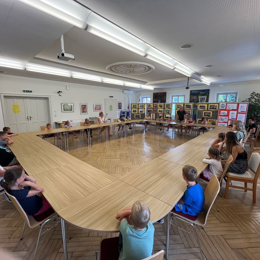 Eine Gruppe von Kindern und Erwachsenen sitzt um einen großen Holztisch in einem Raum mit Holzböden und weißen Wänden.
