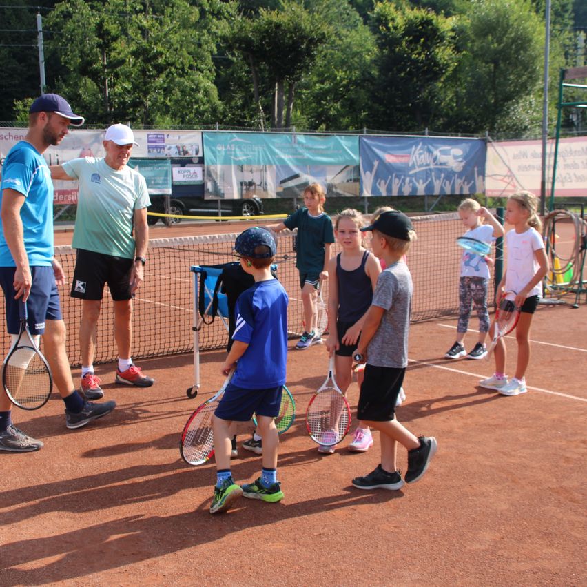 Eine Gruppe von Kindern und zwei Erwachsenen sind auf einem Tennisplatz, wobei ein Erwachsener einen Tennisschläger hält und mit den Kindern spricht.