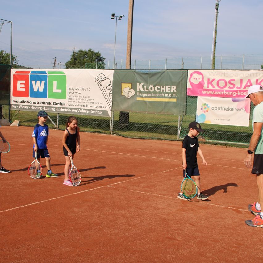 Vier Kinder stehen auf einem Tennisplatz und halten Tennisschläger. Ein Mann in einer weißen Kappe steht hinter ihnen. Werbebanner hängen hinter ihnen am Zaun.