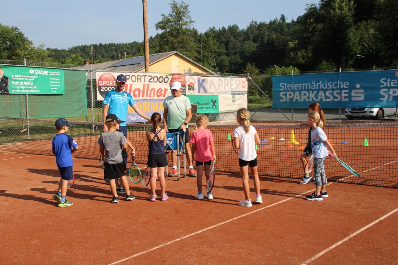 Eine Gruppe von Kindern und zwei Erwachsenen trainiert auf einem Tennisplatz mit Tennisschlägern. Der Platz ist von einem Zaun mit Bannern umgeben. Bäume und ein Gebäude sind im Hintergrund zu sehen.