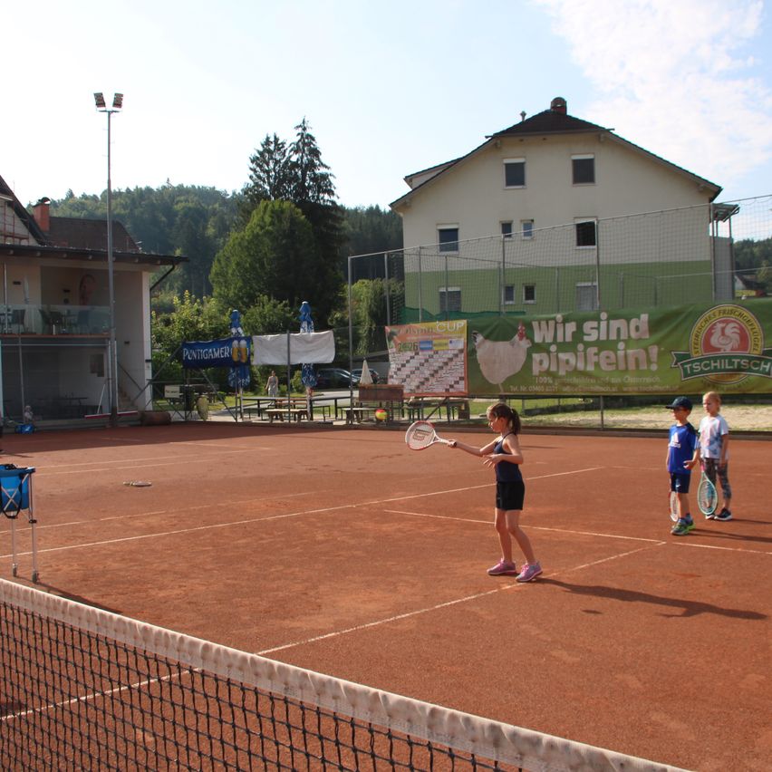 Ein junges Mädchen spielt Tennis auf einem Sandplatz, zwei Jungen beobachten, ein Haus und ein Banner im Hintergrund.