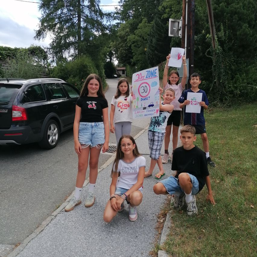 Eine Gruppe von Kindern hält Schilder hoch, die für eine 30 km/h Geschwindigkeitsbegrenzung in ihrer Nachbarschaft werben. Sie stehen auf einem Bürgersteig, neben dem ein schwarzer SUV geparkt ist.
