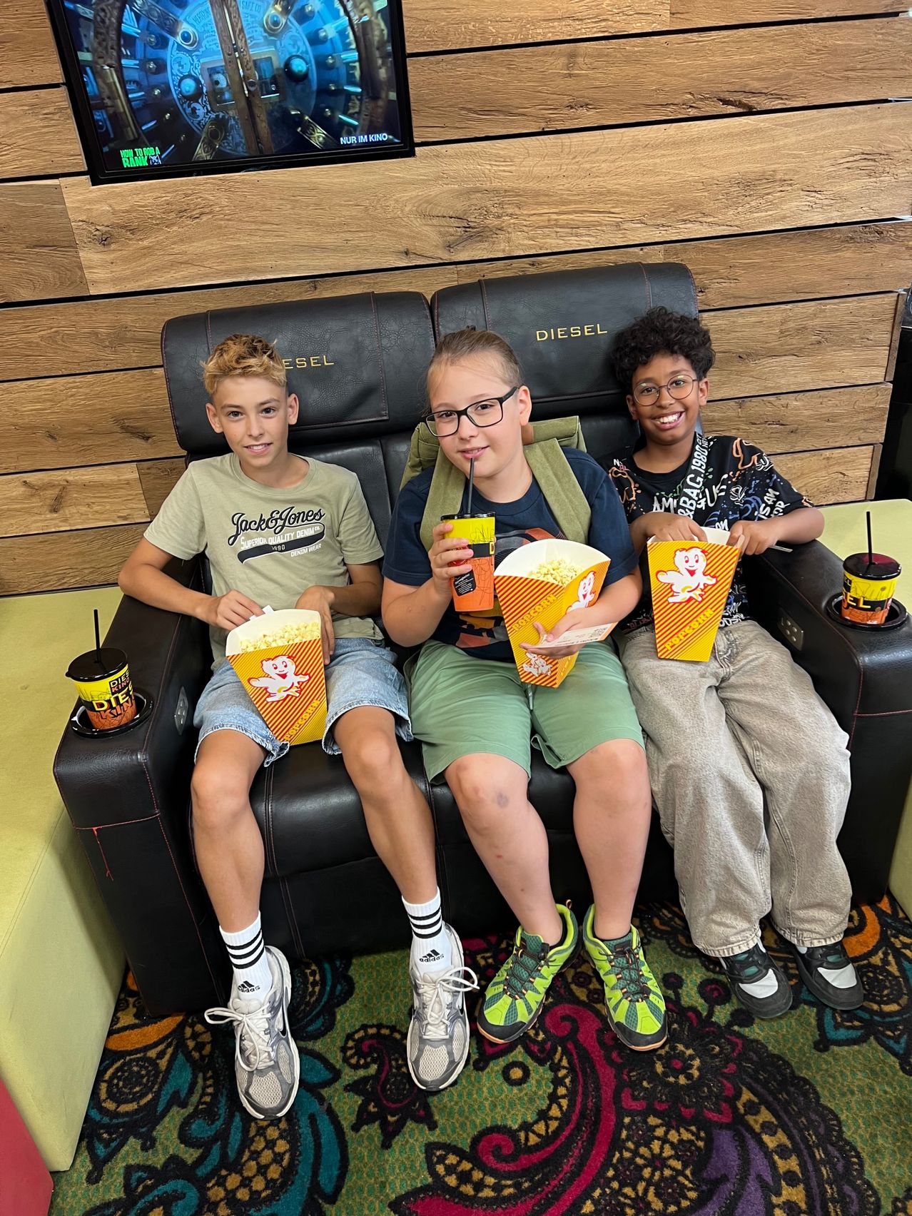Three boys sit on a black theater seat, holding popcorn and drinks. The boy on the left wears a Jack & Jones shirt, the middle one wears glasses and a backpack, and the boy on the right smiles.