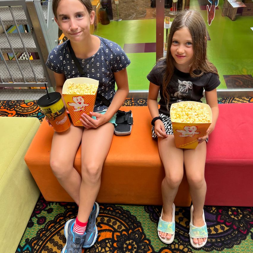 Two young girls are sitting on an orange bench, holding popcorn boxes and smiling. One girl has a cup with a straw. They are in a colorful indoor area with a staircase in the background.