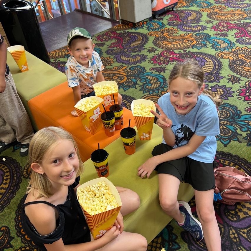 Three children sit on a bench, smiling and posing for a photo. They have popcorn and drinks in front of them. The background features a colorful carpet.