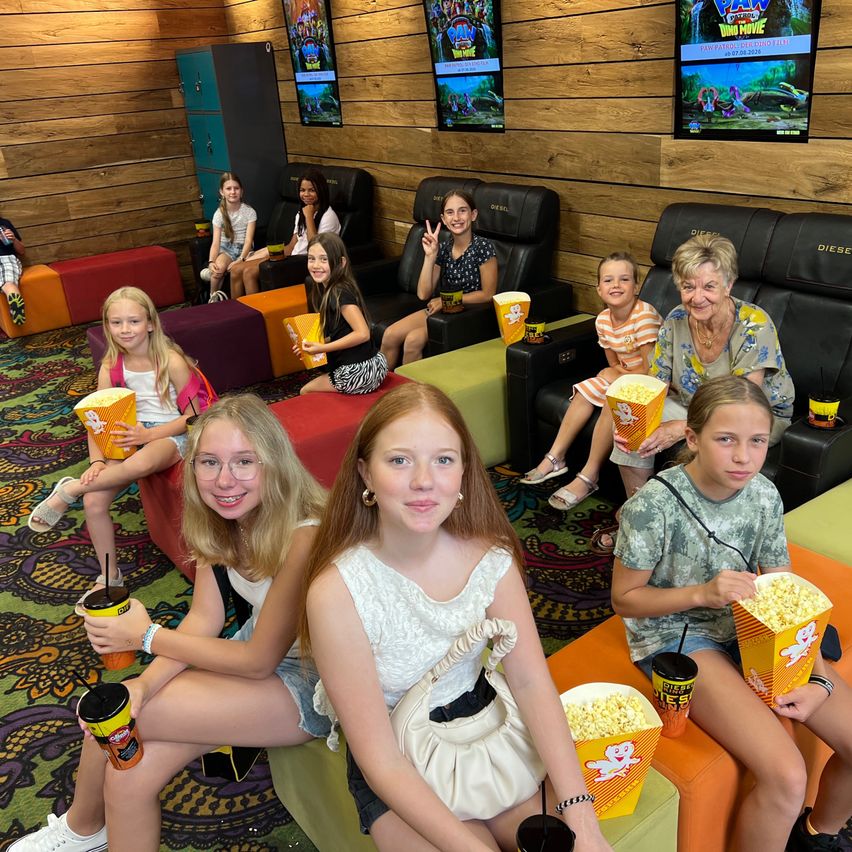 A group of children and adults are sitting in a theater, holding popcorn and drinks. They are smiling and posing for a photo. The theater has a wooden wall and a colorful floor. There are monitors on the wall.