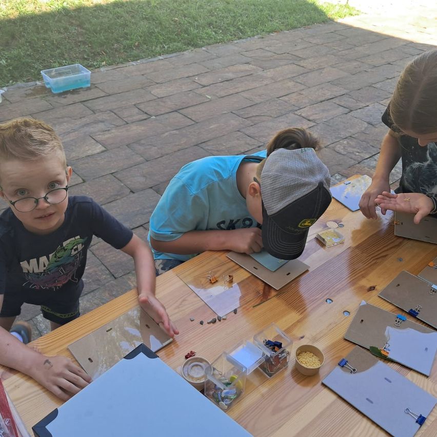 Drei Kinder arbeiten im Freien an einem Holztisch an einem Projekt. Ein Kind trägt eine Brille und ein schwarzes T-Shirt mit einem grafischen Design. Ein weiteres Kind trägt ein blaues T-Shirt und eine Kappe. Das dritte Kind trägt eine Armbanduhr und hält ein Stück Papier. Der Tisch ist mit verschiedenen Materialien und Werkzeugen bestückt.