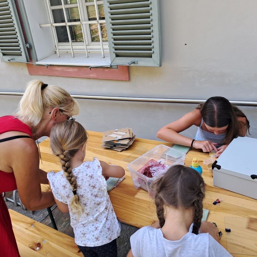 Drei Frauen und ein junges Mädchen sitzen um einen Tisch herum und arbeiten an einem Bastelprojekt. Die Frau in Rot hilft dem Mädchen mit geflochtenen Haaren. Ein weiteres Mädchen sitzt und konzentriert sich auf ihre Aufgabe. Ein Aufbewahrungsbox und ein Plastikbehälter sind auf dem Tisch.