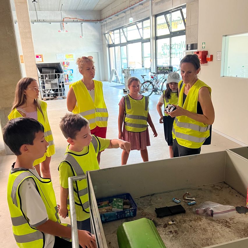 Eine Gruppe von Kindern und Erwachsenen in gelben Sicherheitswesten versammelt sich um einen Tisch mit verschiedenen Gegenständen. Eine Frau in der Mitte hält ein kleines elektronisches Gerät. Hinter ihnen sind Fahrräder in der Nähe einer Glaswand geparkt.