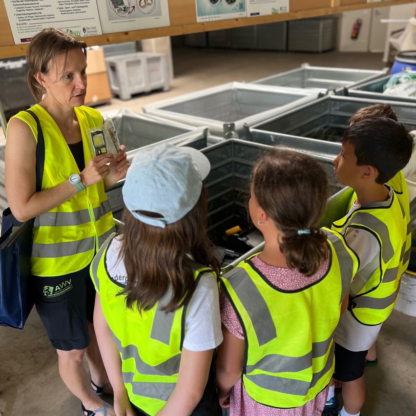 Eine Frau in einer gelben Sicherheitsweste zeigt einer Gruppe von Kindern, die ebenfalls Sicherheitswesten tragen, ein Gerät in einer Lagerhalle.