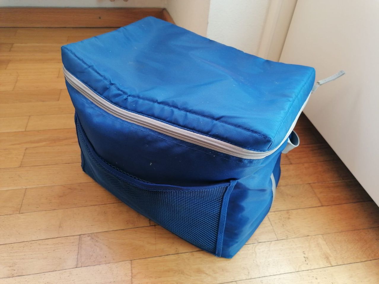 A blue cooler bag with a zipper and mesh pocket is placed on a wooden floor.