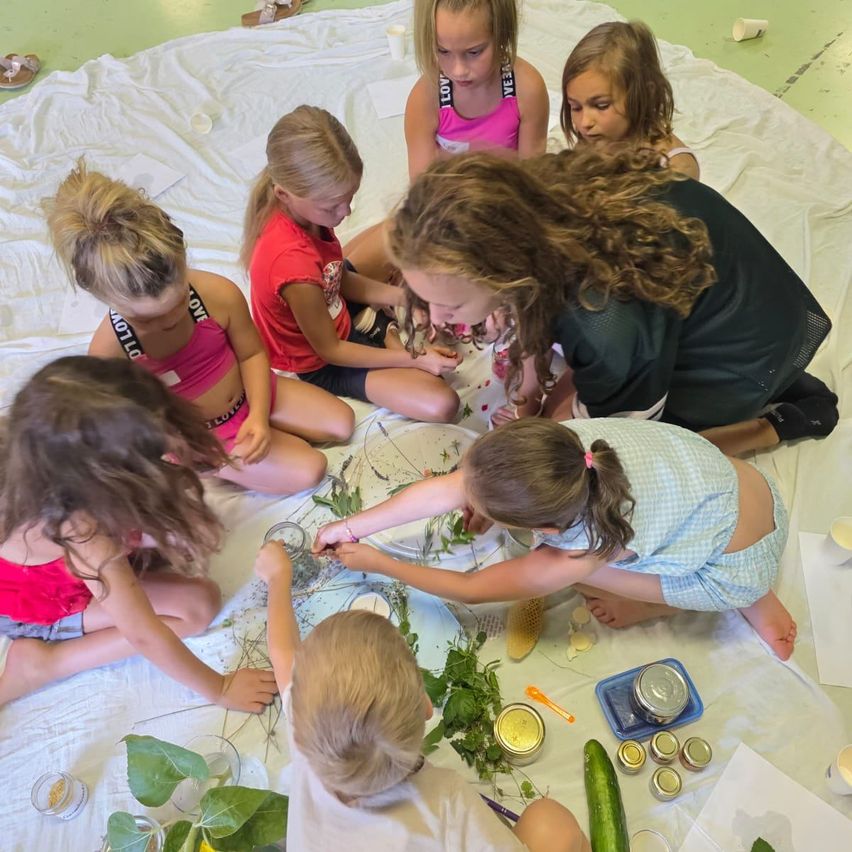 Eine Gruppe junger Mädchen und eine Frau sitzen auf einem weißen Tuch und sind in einer Bastelaktivität verwickelt. Sie sind von verschiedenen Materialien umgeben, darunter Kräuter, Gläser und eine Gurke. Die Frau leitet die Kinder.