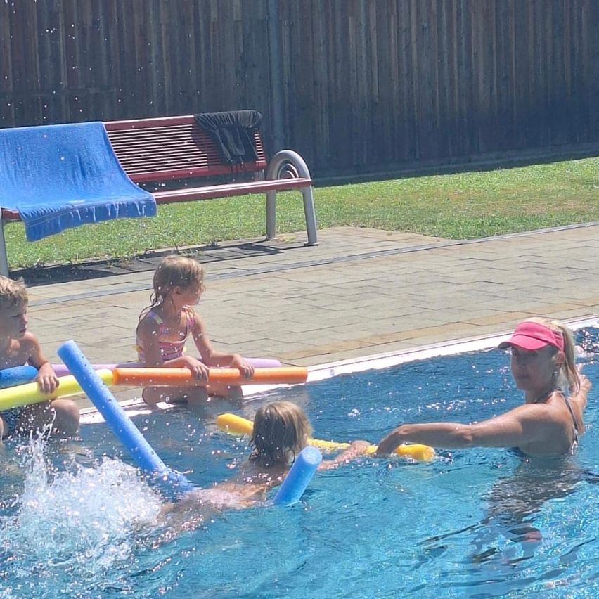 Eine Familie genießt einen Tag am Pool. Eine Frau in einer pinken Kappe schwimmt mit zwei Kindern, während ein Mädchen auf einem Schwimmfloß sitzt.