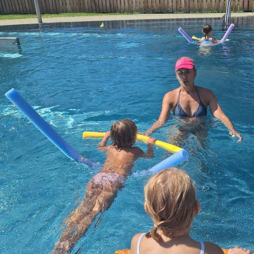 Eine Frau in einer pinken Kappe und einem blauen Bikini schwimmt in einem Pool mit zwei Kindern, eines hält einen gelben Schwimmring. Ein weiteres Kind schwimmt im Hintergrund.