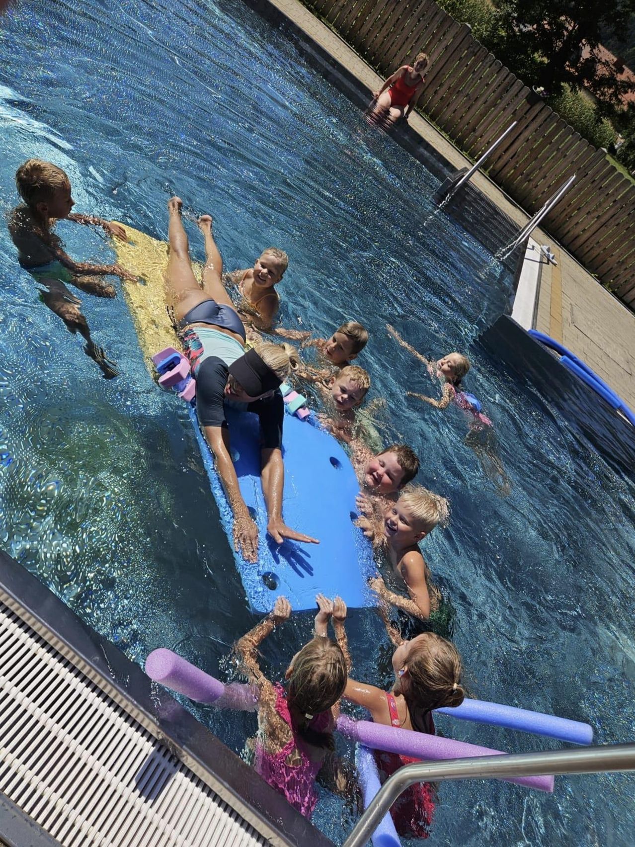 Eine Gruppe von Kindern und Erwachsenen schwimmt in einem Pool, wobei sich ein Erwachsener auf einem blauen Schwimmbrett befindet, während andere es festhalten.