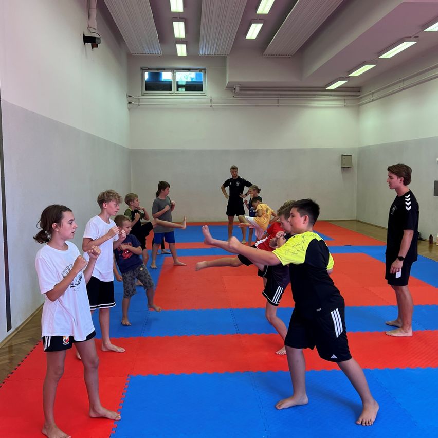 Eine Gruppe von Kindern übt Kampfsport in einer Turnhalle mit einer blau-roten Matte, unter Aufsicht von zwei Erwachsenen.