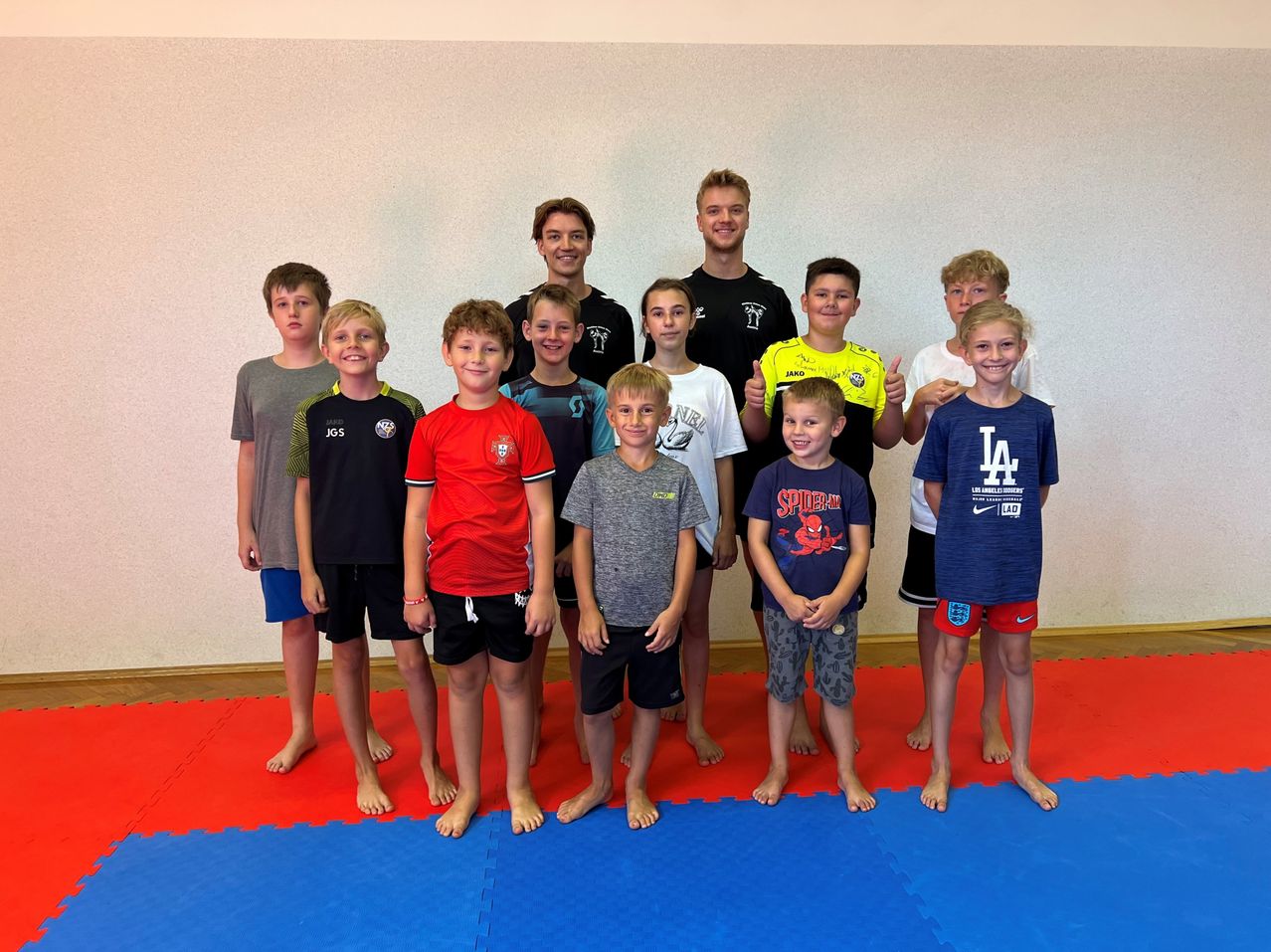 Eine Gruppe junger Jungen und zwei Männer stehen auf einem blauen und roten Teppich und lächeln für ein Foto. Sie tragen verschiedene farbige T-Shirts und Shorts.