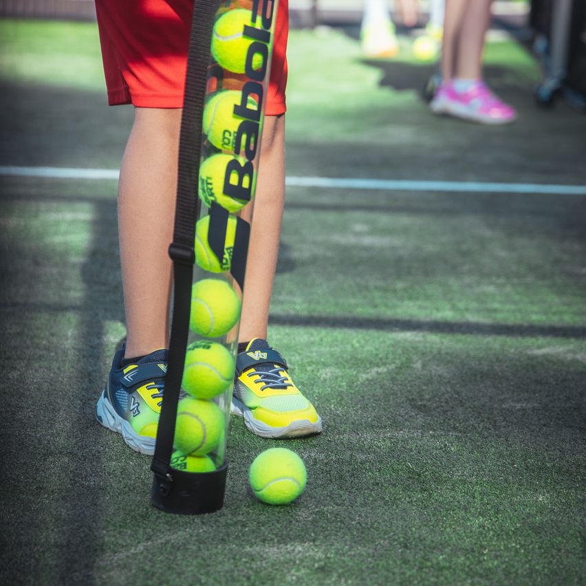 Ein Tennisspieler hält eine Tube mit Tennisbällen, wobei ein Ball auf dem Boden liegt, auf einem Tennisplatz.