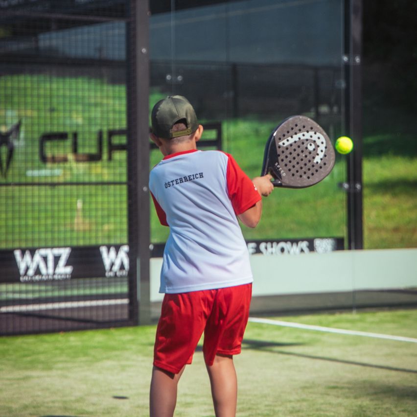 Ein junger Junge mit einem roten und weißen Shirt mit dem Wort 'Osterrech' spielt Tennis auf einem Platz. Er hält einen Tennisschläger und scheint den Ball zu schlagen.