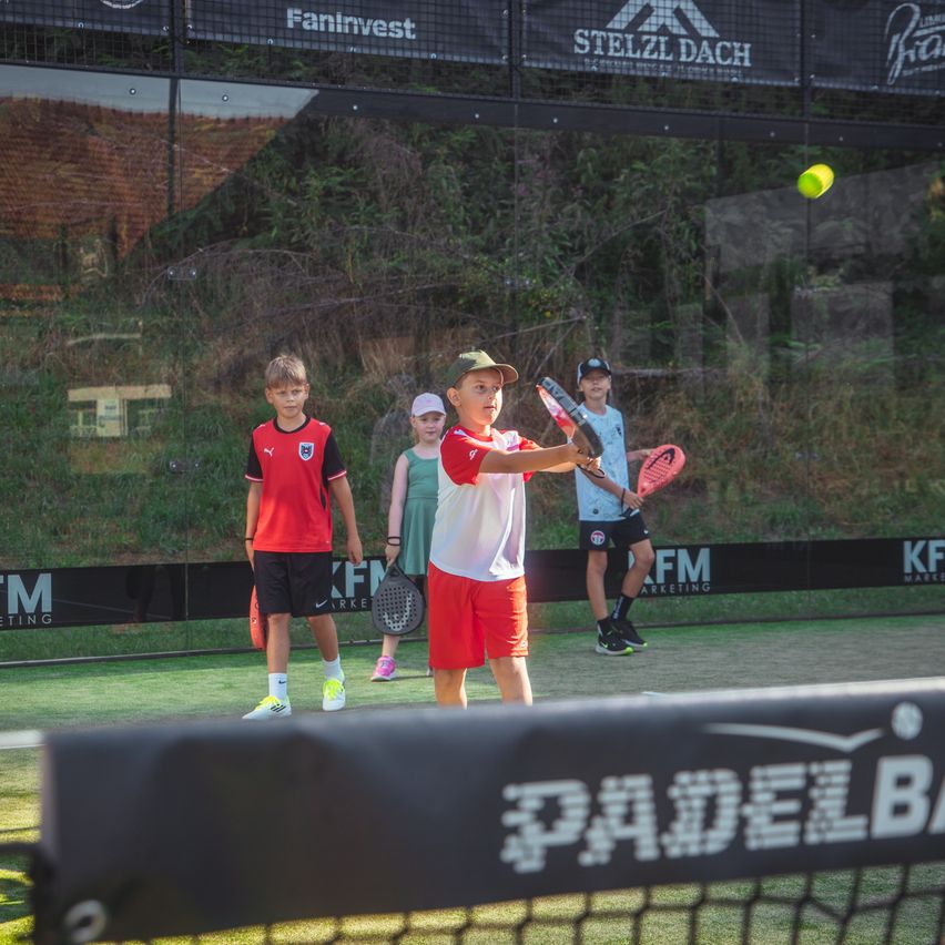 Vier Kinder spielen Paddeltennis auf einem Außenplatz, wobei ein Junge einen gelben Ball schlägt. Der Platz ist von einem Zaun mit Werbung umgeben.