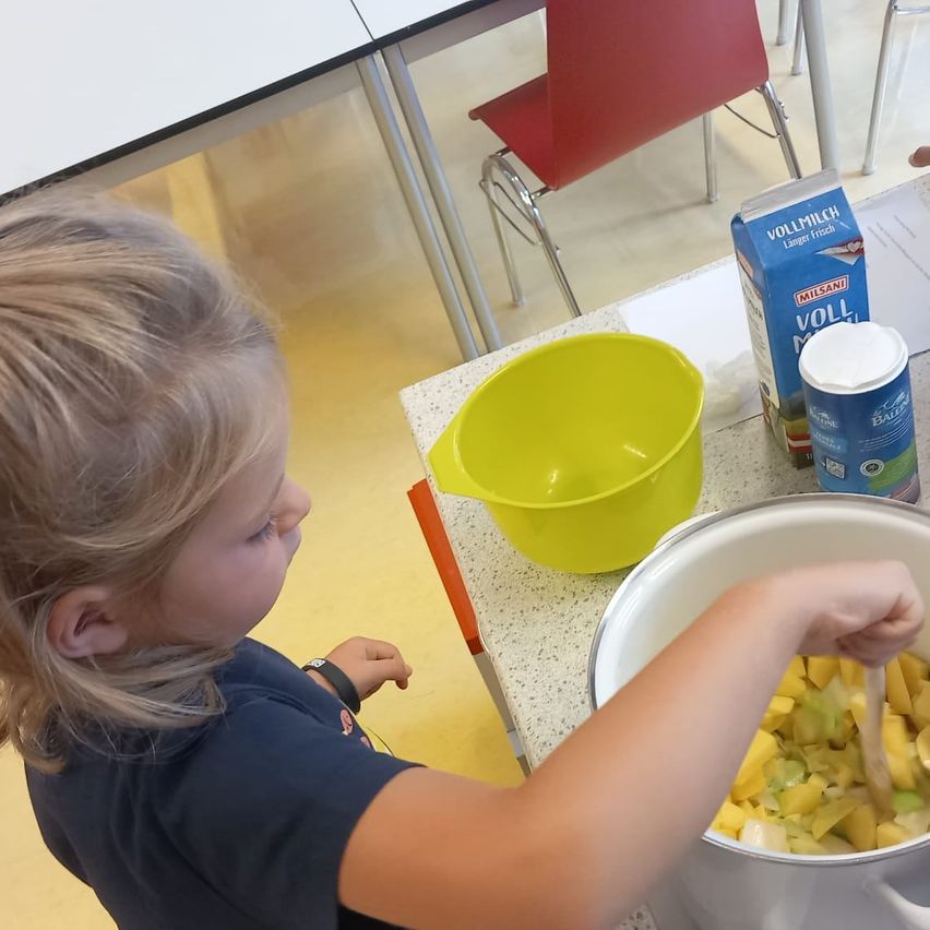 Ein junges Mädchen rührt einen Topf mit Essen mit einem Holzlöffel um. Ein gelber Behälter und ein Milchkarton stehen auf dem Tisch.