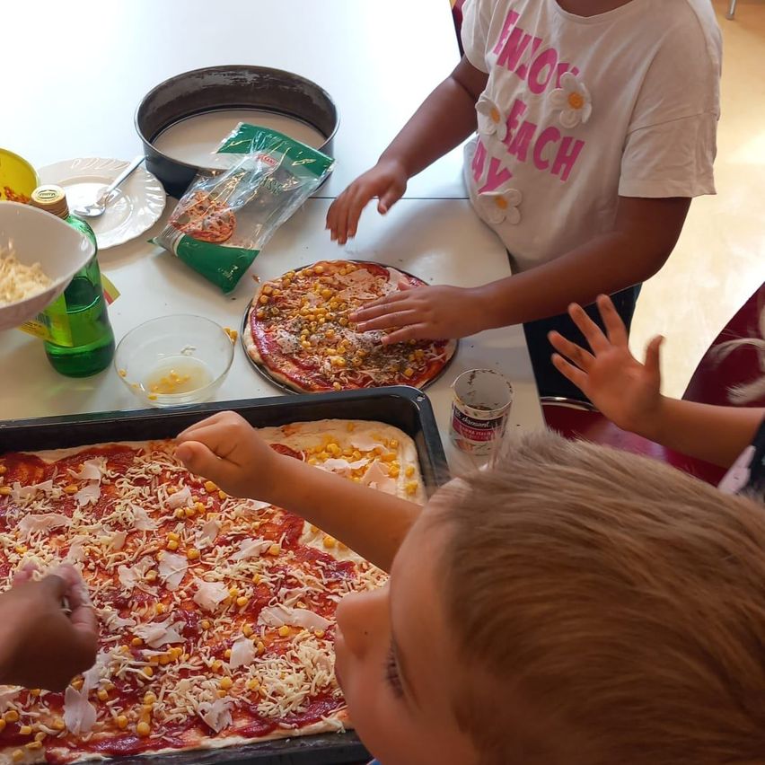 Drei Kinder machen Pizzas mit Toppings wie Käse, Mais und Peperoni auf einem Tisch. Ein Kind fügt Käse zu einer Pizza hinzu.
