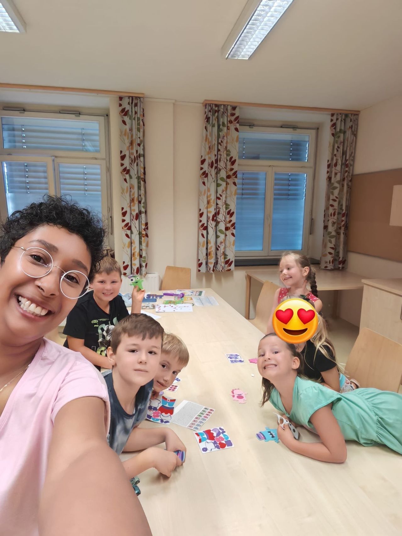 Eine Gruppe von Kindern und einer Frau sitzen an einem Tisch, lächeln und basteln. Die Frau trägt eine Brille und eine Halskette. Die Kinder sind mit Stickern und Papieren beschäftigt.