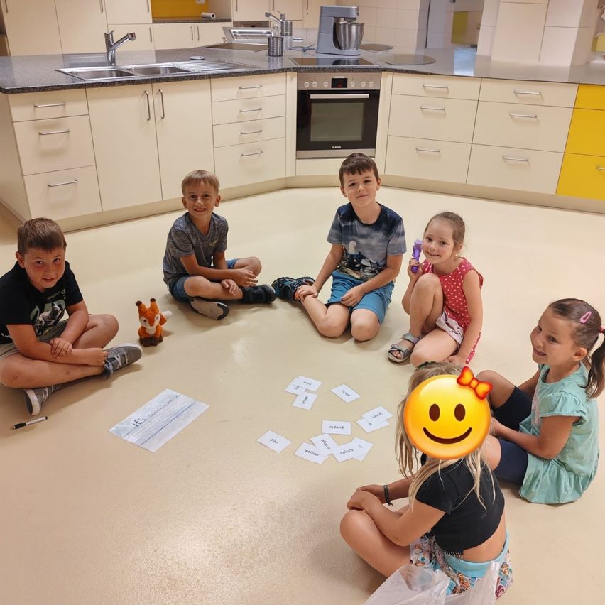 Fünf Kinder sitzen in einem Kreis auf dem Boden in einer Küche, mit Karten und einem Spielzeugfuchs in der Nähe. Ein Mädchen hat einen Smiley-Aufkleber auf dem Kopf.