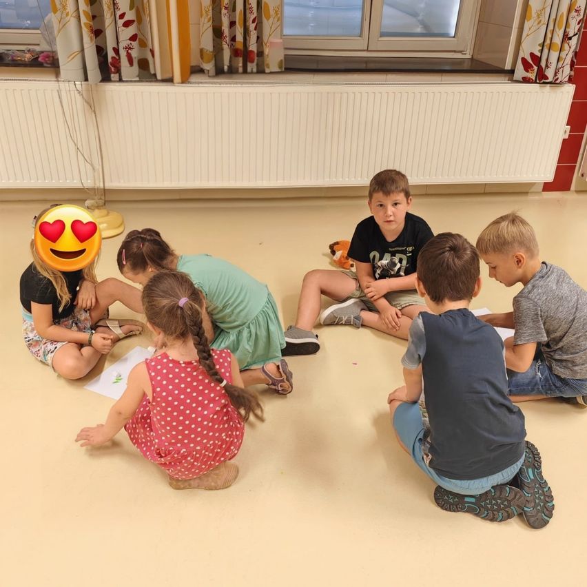 Eine Gruppe von Kindern sitzt auf dem Boden in einem Klassenzimmer und ist in einer Aktivität vertieft. Einige schauen auf Papiere, während andere sich auf etwas anderes konzentrieren.